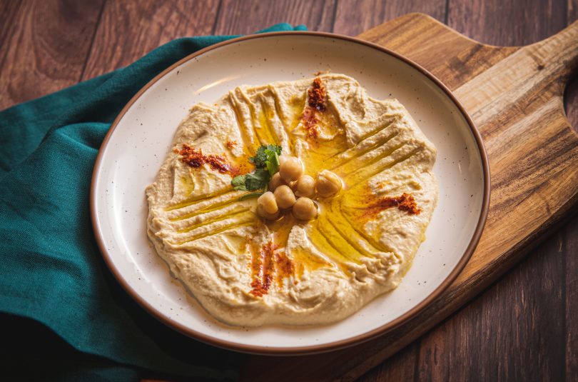 Tan plate with brown rim on wooden table with hummus, oil, and chickpeas. Set on a teal napkin layered on a wooden cutting board and a dark wood table.