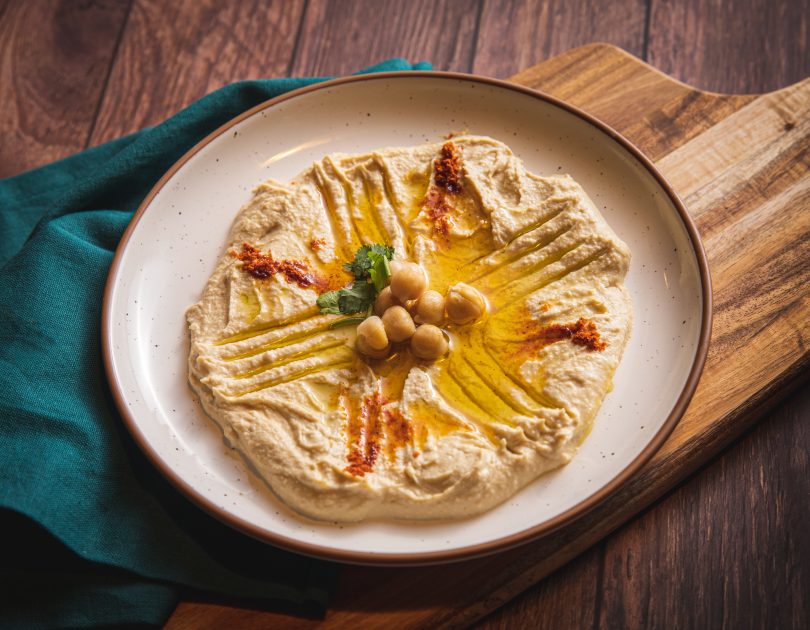 Tan plate with brown rim on wooden table with hummus, oil, and chickpeas. Set on a teal napkin layered on a wooden cutting board and a dark wood table.