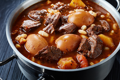Beef Cholent close-up of Traditional Jewish Cholent Hamin - main dish for the Shabbat meal, slow cooked beef with potato, beans and brown eggs in a pot on a black wooden table, vertical view from above.
