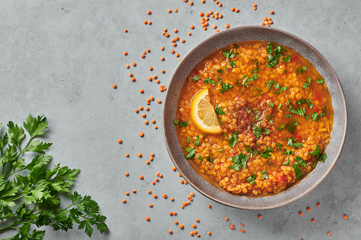 Grey bowl of saucy red lentils with seasonings on a grey background with dry lentils and parsley on a table.