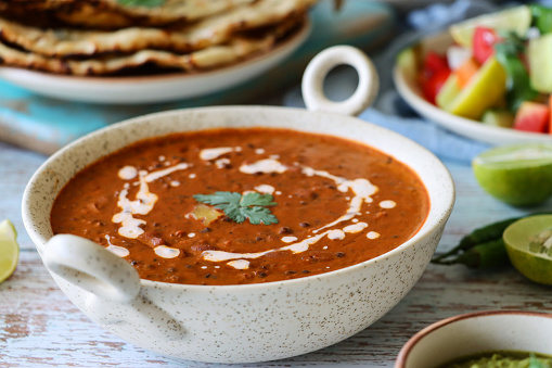 Red soup with white cream drizzle in a white, decorative bowl. Set on a rustic white table with blurred food items in the background including lime, peppers, flat bread, sauce.