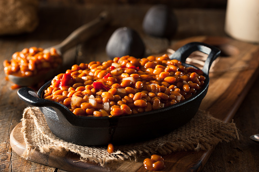 Homemade Barbecue Baked Beans in a Black Skillet Horizontal view of a black skillet of baked beans on a wooden table layered with burlap and a wooden board.