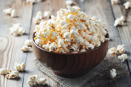 Wooden bowl of popcorn on wooden table with a burlap cloth under the bowl. Popped popcorn kernels are scattered on the table.