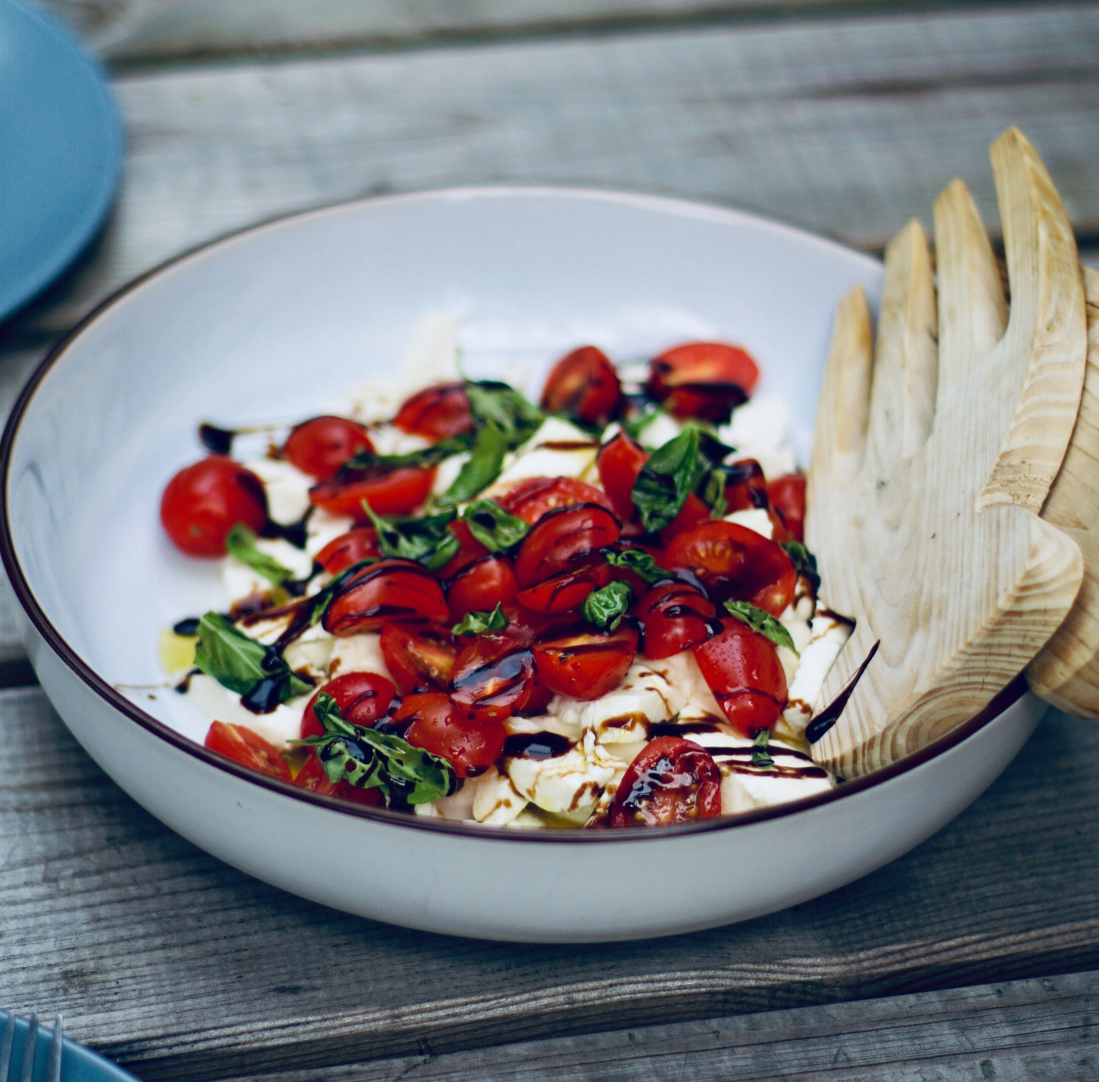 caprese-white-beans A grey wooden table with a white bowl filled with a tomato, cheese, basil, and balsamic salad on it. There are wooden salad claws for serving.