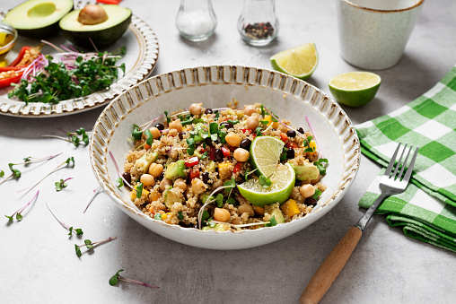 A light grey table with a setting for mealtime including a plate of tomato, halved avocado, onion, and herbs in the background, a salt and pepper shaker set, and a white bowl of quinoa, chickpeas, black beans, and herbs.