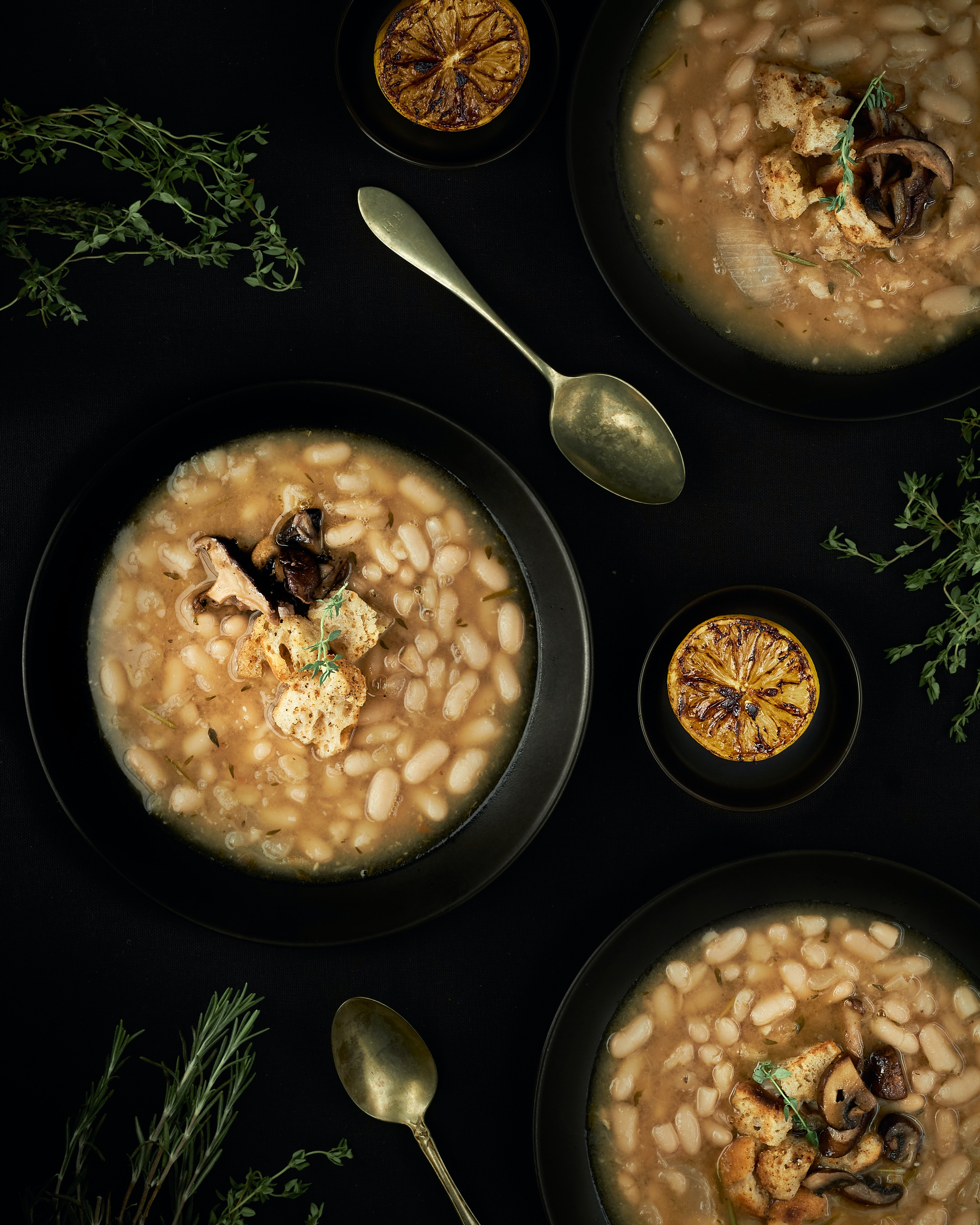 Three settings of black bowls filled with broth and white beans with mushrooms and croutons. Herbs, lemons, and spoons provide decoration on the table.