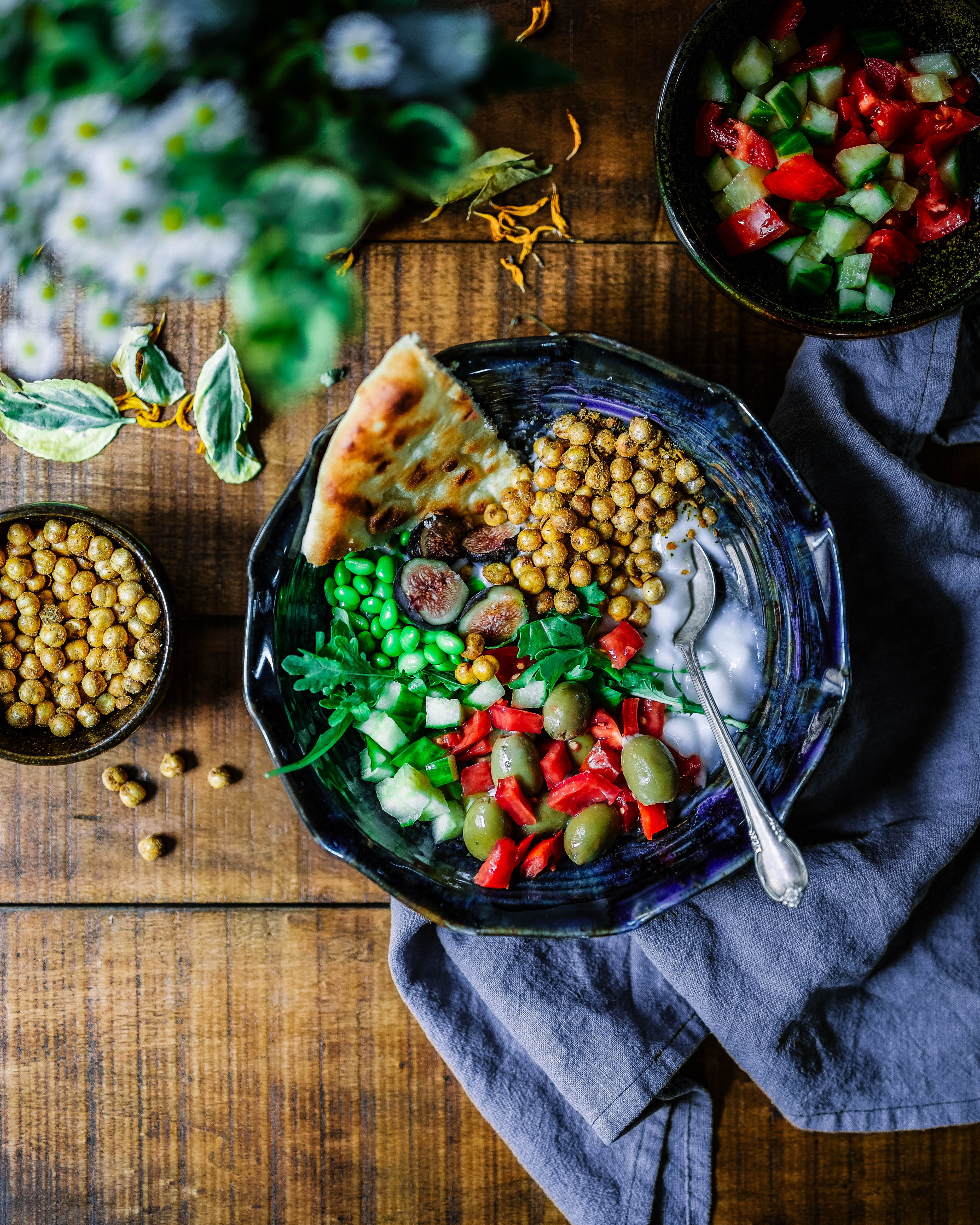 Blue bowl of chickpeas, peppers, olives, soybeans, and cucumber with pita on a wooden table with a bowl of tomato and cucumber as well as an additional bowl of chickpeas.